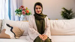 Smiling woman wearing a beige abaya and olive green hijab sitting on a white sofa indoors.