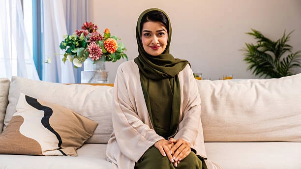 Smiling woman wearing a beige abaya and olive green hijab sitting on a white sofa indoors.